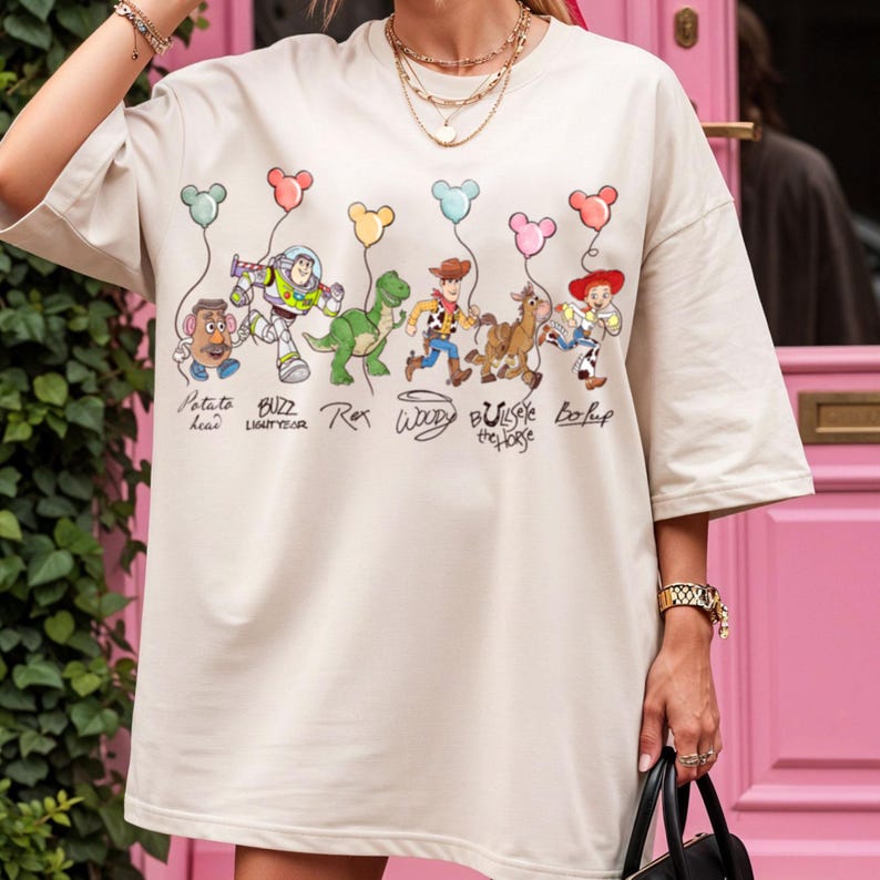May include: Beige oversized t-shirt with a colourful graphic of Toy Story characters holding balloon strings. Characters include Mr. Potato Head, Buzz Lightyear, Rex, Woody, Bullseye the Horse, and Bo Peep. The shirt has short sleeves.