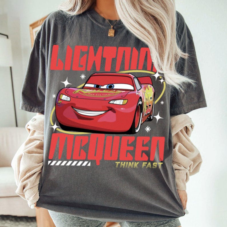 May include: A charcoal gray short-sleeve t-shirt featuring a graphic of the red race car Lightning McQueen. The text reads LIGHTNING MCQUEEN and THINK FAST in bold red and yellow lettering, surrounded by white star accents on the front of the shirt.