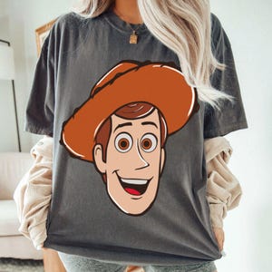 May include: Dark gray t-shirt featuring a cartoon illustration of Woody from Toy Story. The design shows Woody's face with his signature cowboy hat. The shirt is a casual wear item.