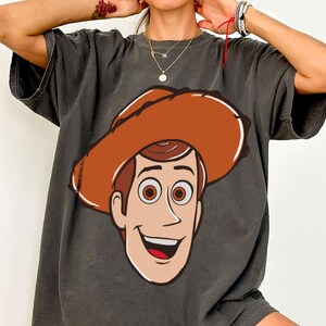 May include: Dark gray t-shirt featuring a cartoon illustration of a smiling cowboy character with a brown hat and hair, and brown eyes. The graphic is centered on the front of the shirt.