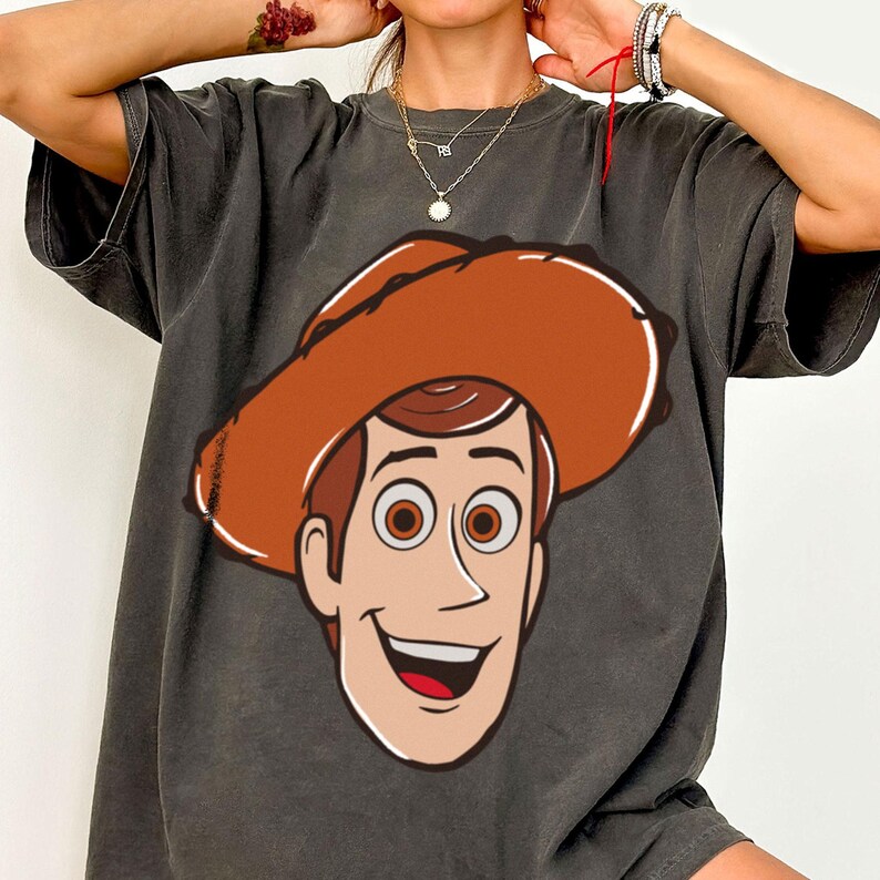 May include: Dark gray t-shirt featuring a cartoon illustration of a smiling cowboy character with a brown hat and hair, and brown eyes. The graphic is centered on the front of the shirt.