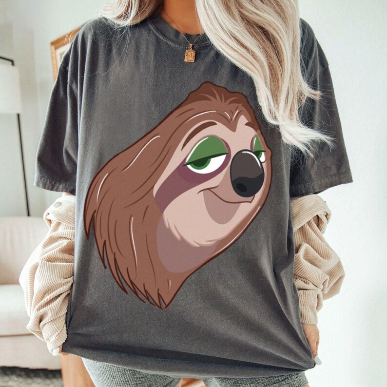 May include: Dark gray t-shirt featuring a cartoon sloth face with brown fur, green eyes, and a black nose. The sloth is smiling. The shirt is a casual style.