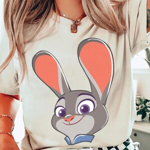 Puede incluir: Una camiseta de color claro con una imagen de dibujos animados de Judy Hopps de Zootopia. La cara del personaje est&aacute; centrada, con grandes orejas con forro rosa, ojos morados y un cuello azul. El dise&ntilde;o est&aacute; impreso en una camiseta de tono neutro.