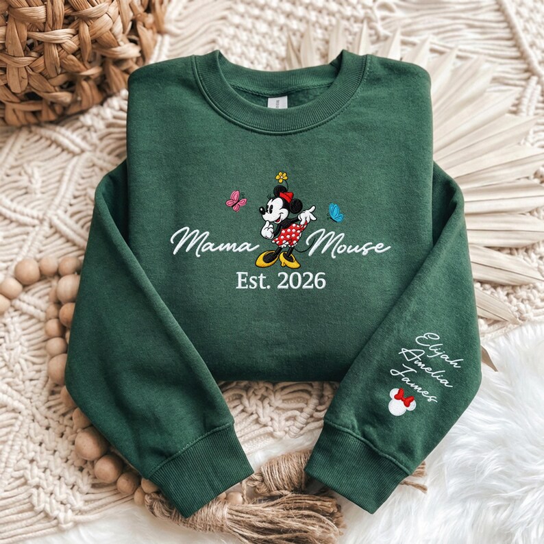 K&ouml;nnte beinhalten: Ein dunkelgr&uuml;nes Sweatshirt mit dem Text "Mama Mouse", einer Minnie Mouse-Grafik und dem Jahr "Est. 2026". Der &Auml;rmel zeigt die Namen "Elijah, Amelia, James" und ein Minnie Mouse-Symbol.