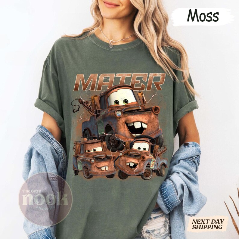 May include: Moss green t-shirt with a graphic of the character Mater from the film Cars. The word "MATER" is printed above the graphic in a distressed, brown font. The Cozy Nook logo is visible on the bottom left.