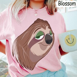 May include: A light pink sweatshirt featuring a large cartoon sloth face with brown fur, green eyes, and a black nose. The word "Blossom" is printed in white in the upper right corner. A white mug with a yellow smiley face is held in the right hand.