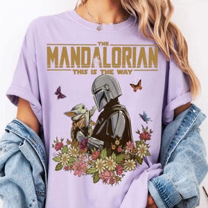 May include: A light purple t-shirt featuring a graphic of The Mandalorian and Grogu surrounded by flowers and butterflies. The text "THE MANDALORIAN" and "THIS IS THE WAY" are printed in gold.