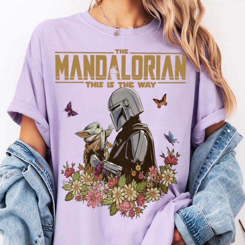 May include: A light purple t-shirt featuring a graphic of The Mandalorian and Grogu surrounded by flowers and butterflies. The text "THE MANDALORIAN" and "THIS IS THE WAY" are printed in gold.
