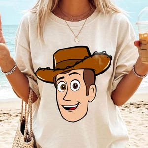 May include: Beige t-shirt featuring a cartoon illustration of a smiling cowboy character with a brown hat and a friendly expression. The design is centered on the shirt, offering a playful and recognizable image.