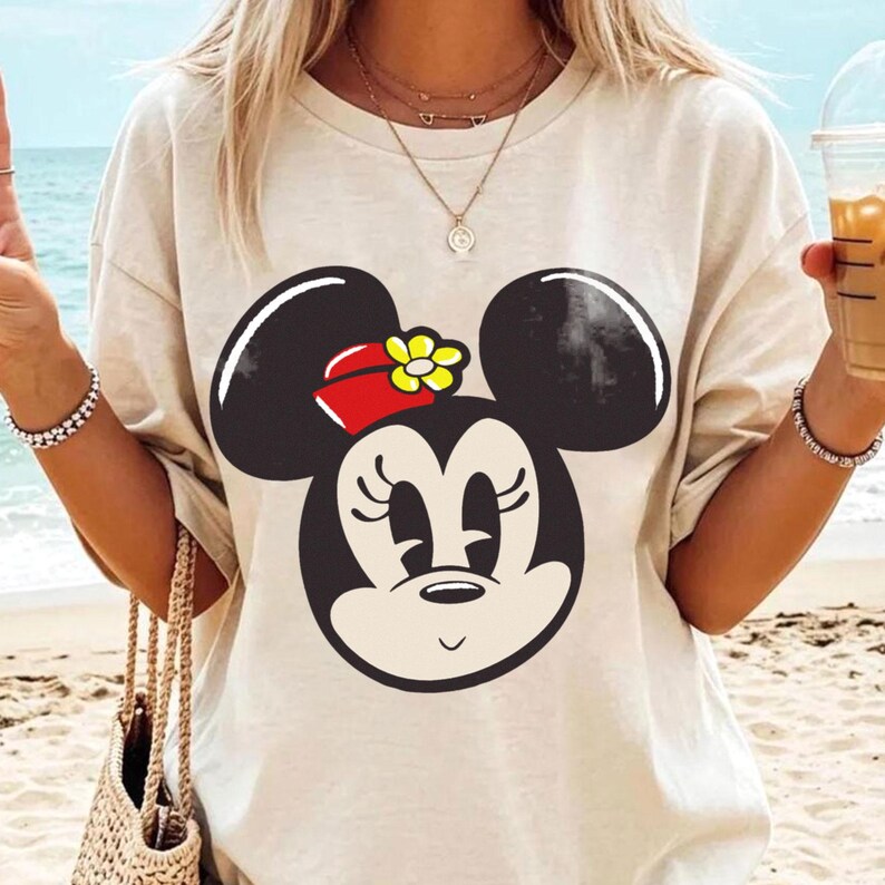 Peut inclure: T-shirt beige avec un grand dessin de Minnie Mouse. Le motif comprend des oreilles noires, un n&oelig;ud rouge avec une fleur jaune et un visage souriant. Le t-shirt est de style d&eacute;contract&eacute;.