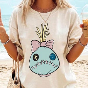 May include: Beige t-shirt featuring a cartoon character design. The design is a light blue sack-like figure with a pink bow, yellow hair, and stitched details. The character has a black cross for an eye and a blue button eye.