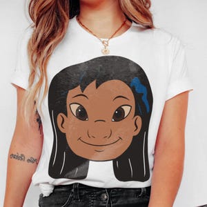 Puede incluir: Camiseta blanca con una ilustraci&oacute;n de dibujos animados de una ni&ntilde;a con cabello oscuro, piel morena y un lazo azul. El gr&aacute;fico est&aacute; centrado en la parte delantera. La camiseta tiene mangas cortas y cuello redondo.
