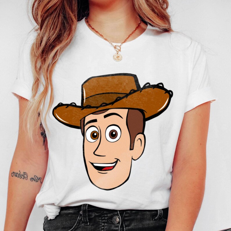 May include: White t-shirt featuring a cartoon illustration of Woody from Toy Story. The character has a brown cowboy hat, brown hair, and a smiling face. The shirt is a classic crew neck style.