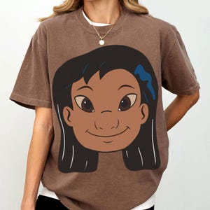 Puede incluir: Camiseta marr&oacute;n con una ilustraci&oacute;n de dibujos animados de la cara de una ni&ntilde;a. La ni&ntilde;a tiene el pelo negro y un accesorio azul para el pelo. El gr&aacute;fico est&aacute; centrado en la parte delantera de la camiseta. La camiseta tiene mangas cortas y cuello redondo.