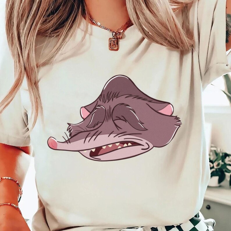Peut inclure: T-shirt cr&egrave;me avec une illustration de dessin anim&eacute; d'une cr&eacute;ature endormie, violet-gris avec des accents roses. La cr&eacute;ature a les yeux ferm&eacute;s, un long museau et des dents visibles. Le t-shirt est un v&ecirc;tement d&eacute;contract&eacute; de tous les jours.