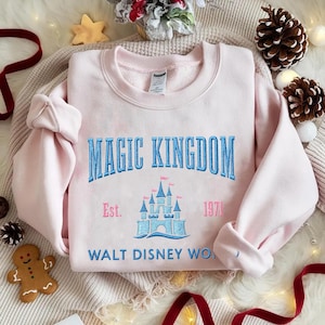 May include: A light pink sweatshirt with the words "MAGIC KINGDOM" embroidered in blue, along with a castle graphic and the text "Est. 1971 WALT DISNEY WO...". The sweatshirt is folded on a cream-colored surface, with holiday decorations.