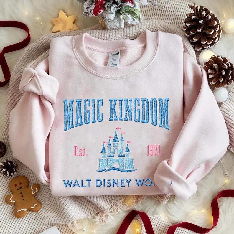 May include: A light pink sweatshirt with the words "MAGIC KINGDOM" embroidered in blue, along with a castle graphic and the text "Est. 1971 WALT DISNEY WO...". The sweatshirt is folded on a cream-colored surface, with holiday decorations.