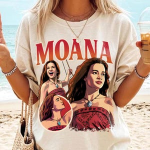 以下が含まれることがあります： 胸元に大きな赤い文字でMOANAとプリントされたベージュの半袖Tシャツ。テキストの下には、赤い柄のトップスと青い石のペンダントネックレスを身に着けたキャラクター、モアナのイラストが3つ描かれています。