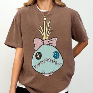 May include: Brown t-shirt featuring a cartoon voodoo doll design. The doll is light blue with a pink bow, yellow hair, and stitched details. The shirt has short sleeves and a crew neck.