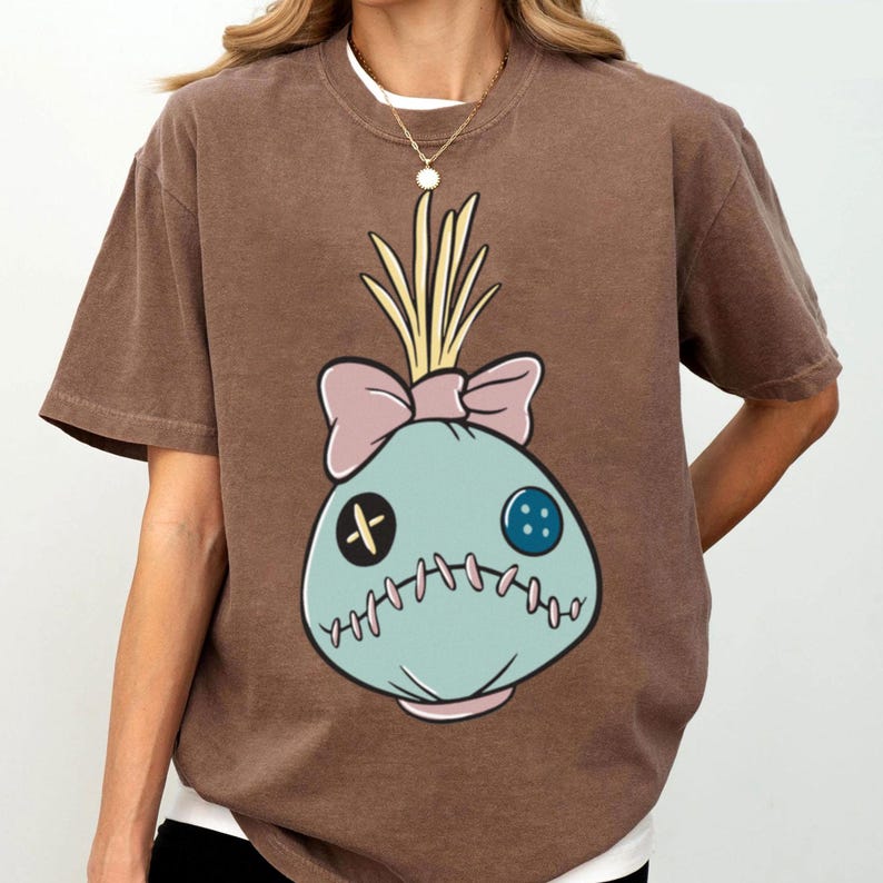 May include: Brown t-shirt featuring a cartoon voodoo doll design. The doll is light blue with a pink bow, yellow hair, and stitched details. The shirt has short sleeves and a crew neck.