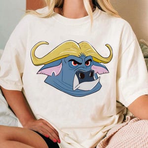 May include: Cream-colored t-shirt featuring a cartoon buffalo head. The buffalo has a blue face, yellow horns, and red eyes. The shirt is a casual style, suitable for everyday wear.