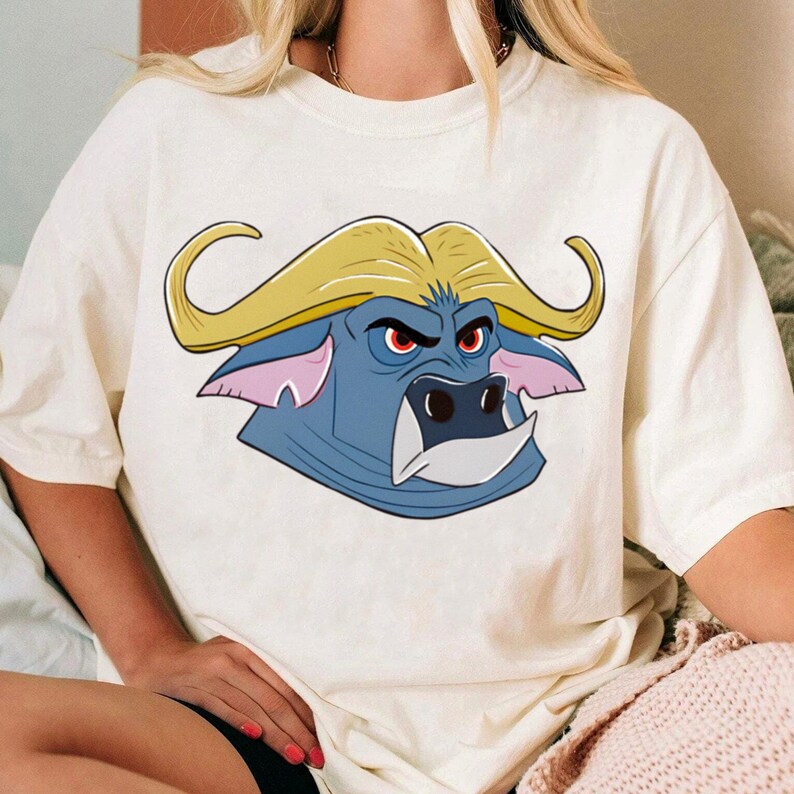 May include: Cream-colored t-shirt featuring a cartoon buffalo head. The buffalo has a blue face, yellow horns, and red eyes. The shirt is a casual style, suitable for everyday wear.