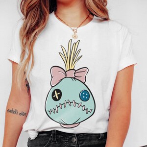 May include: White t-shirt featuring a cartoon-style voodoo doll design. The doll is light blue with a pink bow, yellow hair, and stitched details. It has a black cross and a blue button for eyes.