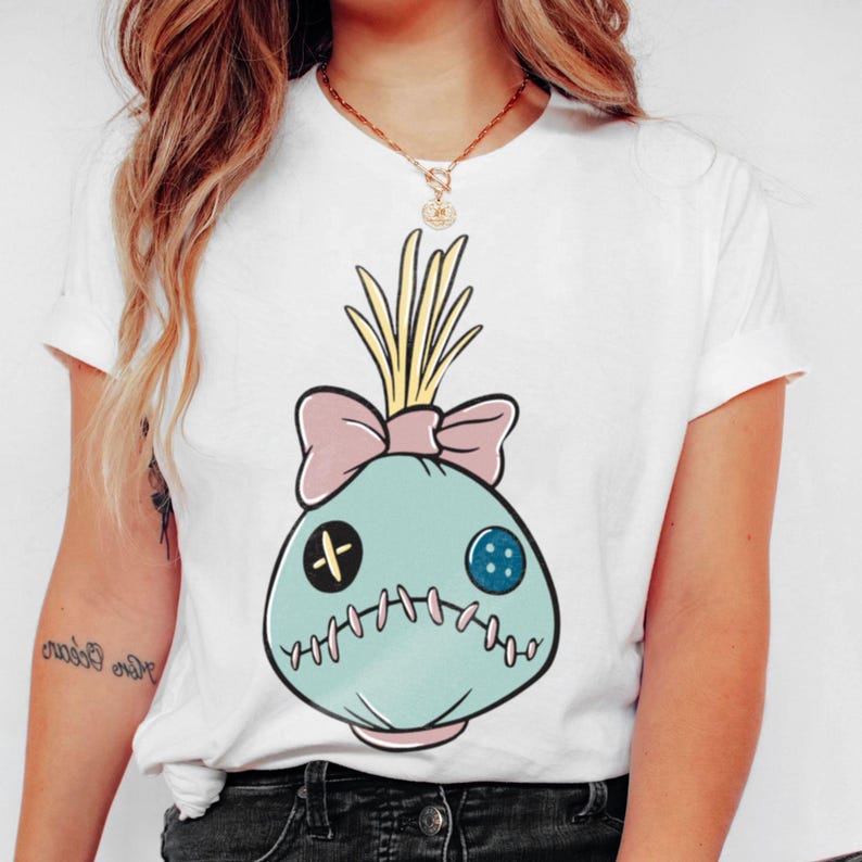 May include: White t-shirt featuring a cartoon-style voodoo doll design. The doll is light blue with a pink bow, yellow hair, and stitched details. It has a black cross and a blue button for eyes.