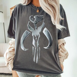 May include: Dark grey t-shirt with a large, metallic silver emblem. The emblem has a stylized, angular design with curved elements. The shirt is a relaxed fit, with short sleeves and a crew neck.