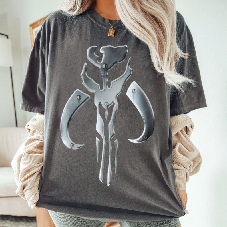 May include: Dark grey t-shirt with a large, metallic silver emblem. The emblem has a stylized, angular design with curved elements. The shirt is a relaxed fit, with short sleeves and a crew neck.