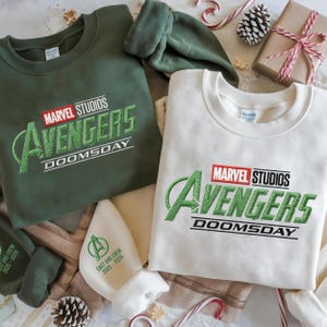 May include: Two sweatshirts with the Marvel Studios Avengers Doomsday logo. One is olive green, the other is off-white. A matching green hat and a white hat with text are also visible. Festive holiday decorations are in the background.
