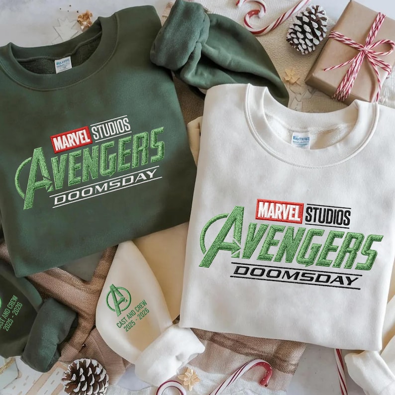 May include: Two sweatshirts with the Marvel Studios Avengers Doomsday logo. One is olive green, the other is off-white. A matching green hat and a white hat with text are also visible. Festive holiday decorations are in the background.