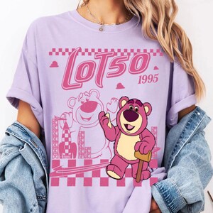 May include: A lilac-colored t-shirt featuring a pink graphic of Lotso, the bear character from Toy Story, with the text "LOTSO 1995". The design includes a checkered pattern and a rocket ship.