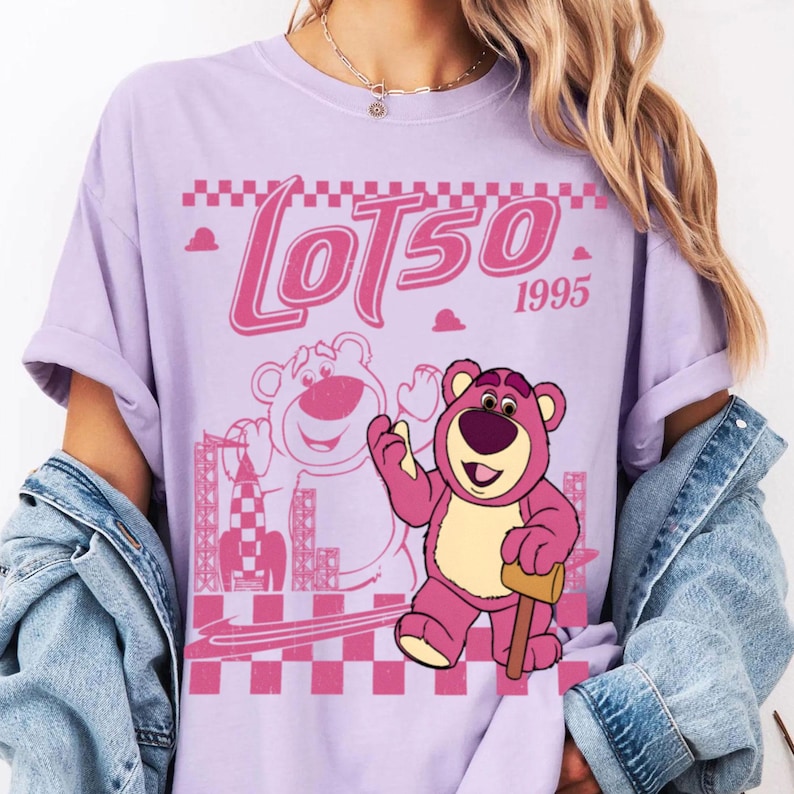 May include: A lilac-colored t-shirt featuring a pink graphic of Lotso, the bear character from Toy Story, with the text "LOTSO 1995". The design includes a checkered pattern and a rocket ship.