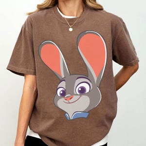 Puede incluir: Camiseta marr&oacute;n con una imagen de dibujos animados de Judy Hopps de Zootopia. El gr&aacute;fico muestra la cara de Judy con orejas grandes, ojos morados y una expresi&oacute;n sonriente. La camiseta tiene mangas cortas y cuello redondo.