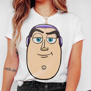 May include: White t-shirt featuring a cartoon illustration of Buzz Lightyear's face. The design includes his signature purple headpiece, blue eyes, and a smiling expression. The shirt is a classic crew neck style.