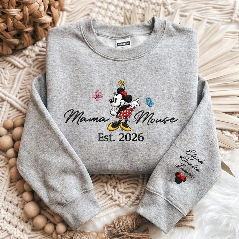 K&ouml;nnte beinhalten: Ein graues Sweatshirt mit den gestickten W&ouml;rtern "Mama Mouse" und "Est. 2026". Das Design umfasst eine Cartoon-Maus, Schmetterlinge und Namen auf dem &Auml;rmel. Ein l&auml;ssiger, bequemer Sweatshirt-Stil.