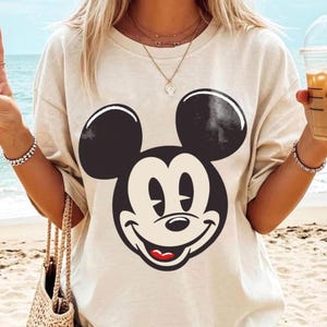 May include: A beige t-shirt featuring a large, smiling Mickey Mouse face in black and white. The classic cartoon character's face is centered on the shirt, with a red mouth and a cream-colored face. The shirt has short sleeves and a relaxed fit.
