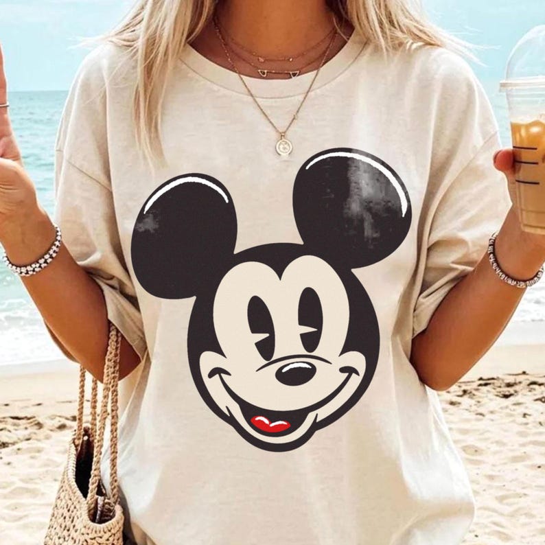 May include: A beige t-shirt featuring a large, smiling Mickey Mouse face in black and white. The classic cartoon character's face is centered on the shirt, with a red mouth and a cream-colored face. The shirt has short sleeves and a relaxed fit.