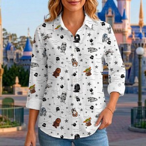 May include: White button-down shirt with a Star Wars pattern. The shirt displays various characters, spaceships, and star graphics in black and grey. The long-sleeved shirt is worn by a person in front of a blurred background.
