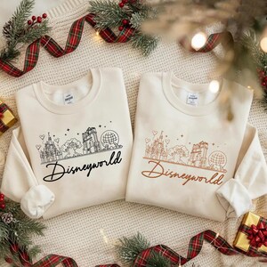 May include: Two cream-colored sweatshirts with long sleeves, each featuring a line drawing of a castle, tree, and globe, with the word "Disneyworld" below. One design is in black, the other in brown.