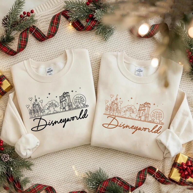 May include: Two cream-colored sweatshirts with long sleeves, each featuring a line drawing of a castle, tree, and globe, with the word "Disneyworld" below. One design is in black, the other in brown.