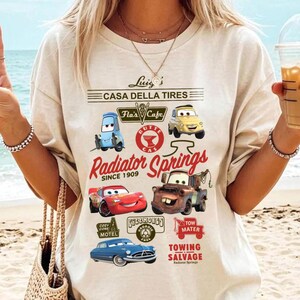 Pu&ograve; includere: Una t-shirt a maniche corte color crema con una stampa grafica di personaggi e luoghi del film Cars. Il design include testi come Luigi's Casa Della Tires, Flo's V8 Cafe, Radiator Springs Since 1909 e Tow Mater Towing Salvage.