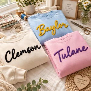May include: Three crewneck sweatshirts in light blue, cream, and light pink. The light blue sweatshirt features the yellow text Baylor, the cream sweatshirt features the black text Clemson, and the light pink sweatshirt features the purple text Tulane.