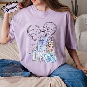 May include: A light purple t-shirt featuring a graphic of Elsa from Frozen, a castle, and fireworks within a Mickey Mouse head outline. The shirt has the text "Comfort Colors Orchid" at the top and "Tito Cozy Nook" at the bottom.