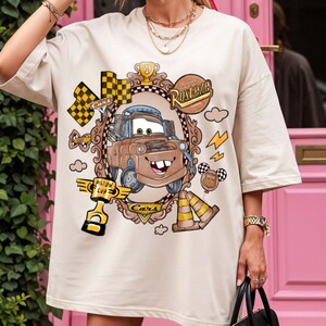 May include: Beige t-shirt featuring a cartoon image of Mater from the movie Cars, framed by a decorative border. The design includes racing flags, lightning bolts, and the words "Rusteze" and "Piston Cup".