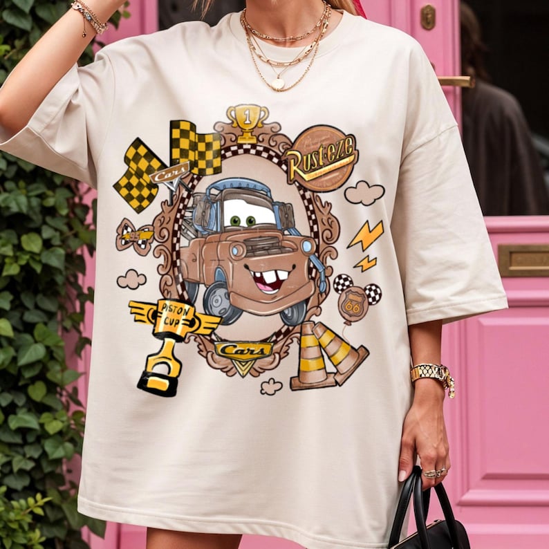 May include: Beige t-shirt featuring a cartoon image of Mater from the movie Cars, framed by a decorative border. The design includes racing flags, lightning bolts, and the words "Rusteze" and "Piston Cup".