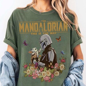 May include: Green t-shirt featuring a graphic of The Mandalorian and Grogu surrounded by flowers and butterflies. The text "THE MANDALORIAN" and "THIS IS THE WAY" are printed in gold across the top.