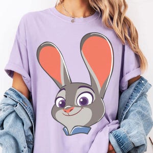 Puede incluir: Camiseta lila claro con un dise&ntilde;o de conejo de dibujos animados. El conejo tiene pelaje gris, orejas grandes con interiores naranjas y ojos morados. Lleva un cuello azul. La camiseta tiene mangas cortas y un corte holgado.