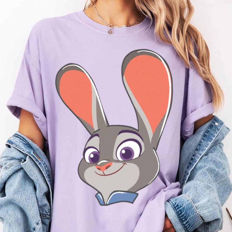 Puede incluir: Camiseta lila claro con un dise&ntilde;o de conejo de dibujos animados. El conejo tiene pelaje gris, orejas grandes con interiores naranjas y ojos morados. Lleva un cuello azul. La camiseta tiene mangas cortas y un corte holgado.